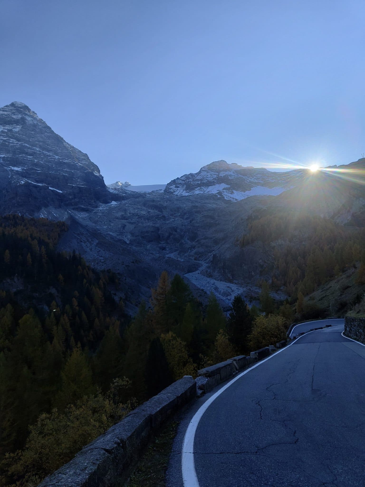 An alpine road curving toward a low sun over the ridge