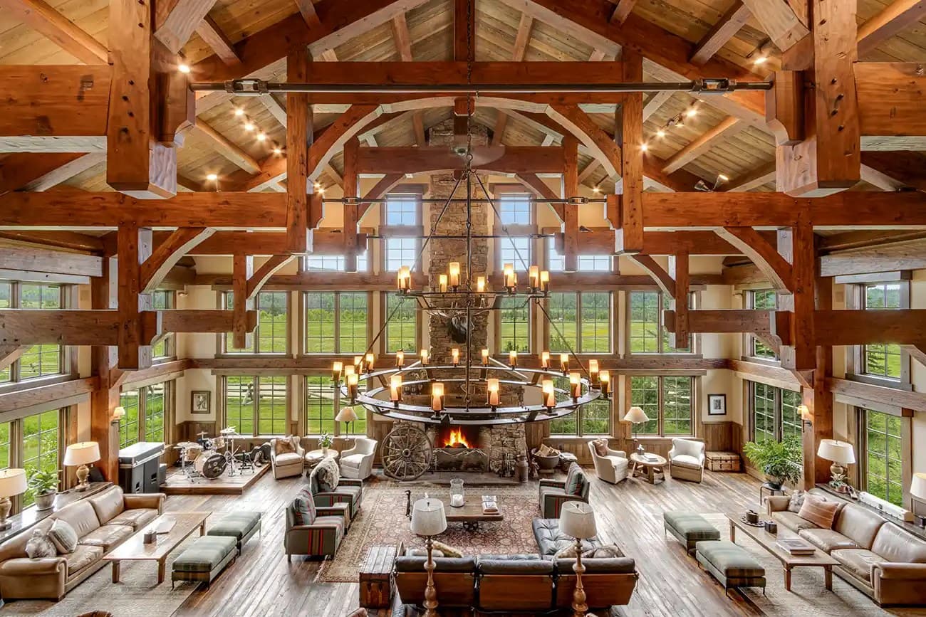 A timber-framed ranch great room with a stone fireplace, iron chandelier, and tall windows onto open pasture