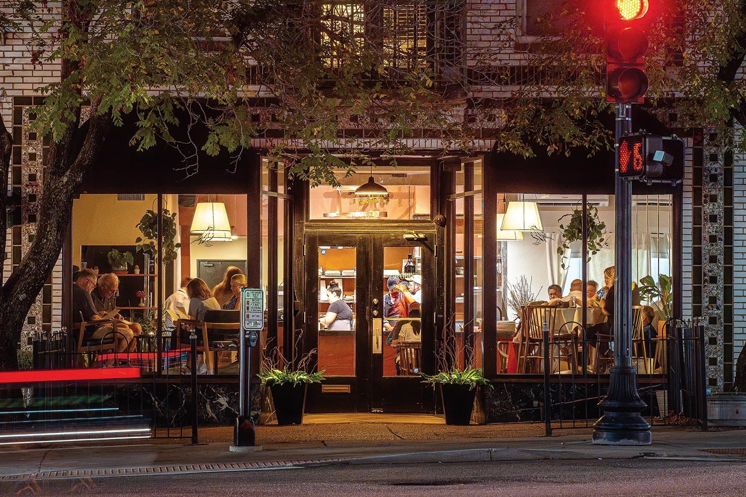 A neighborhood restaurant glowing at night, diners visible through tall windows beneath a red traffic signal